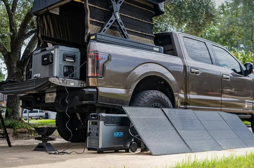 ABOK portable power stations and a foldable solar panel set up beside a truck campsite.